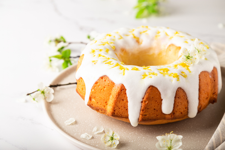Fresh homemade lemon cake decorated with white glaze and zest on white marble background with branches of blossoming plum